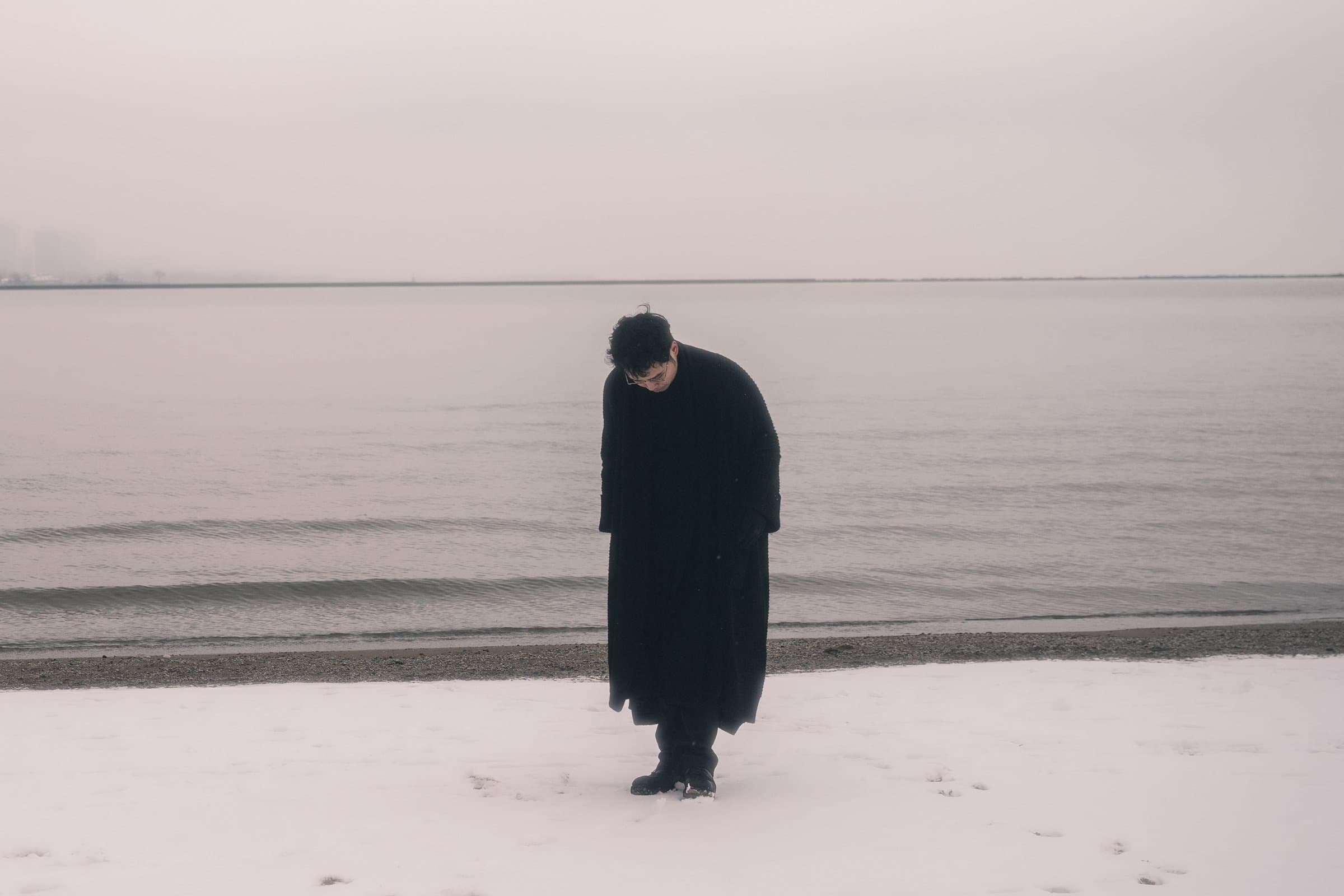 Standing on a snow-covered beach in a long black coat, head bowed, the lake and a pale winter sky behind