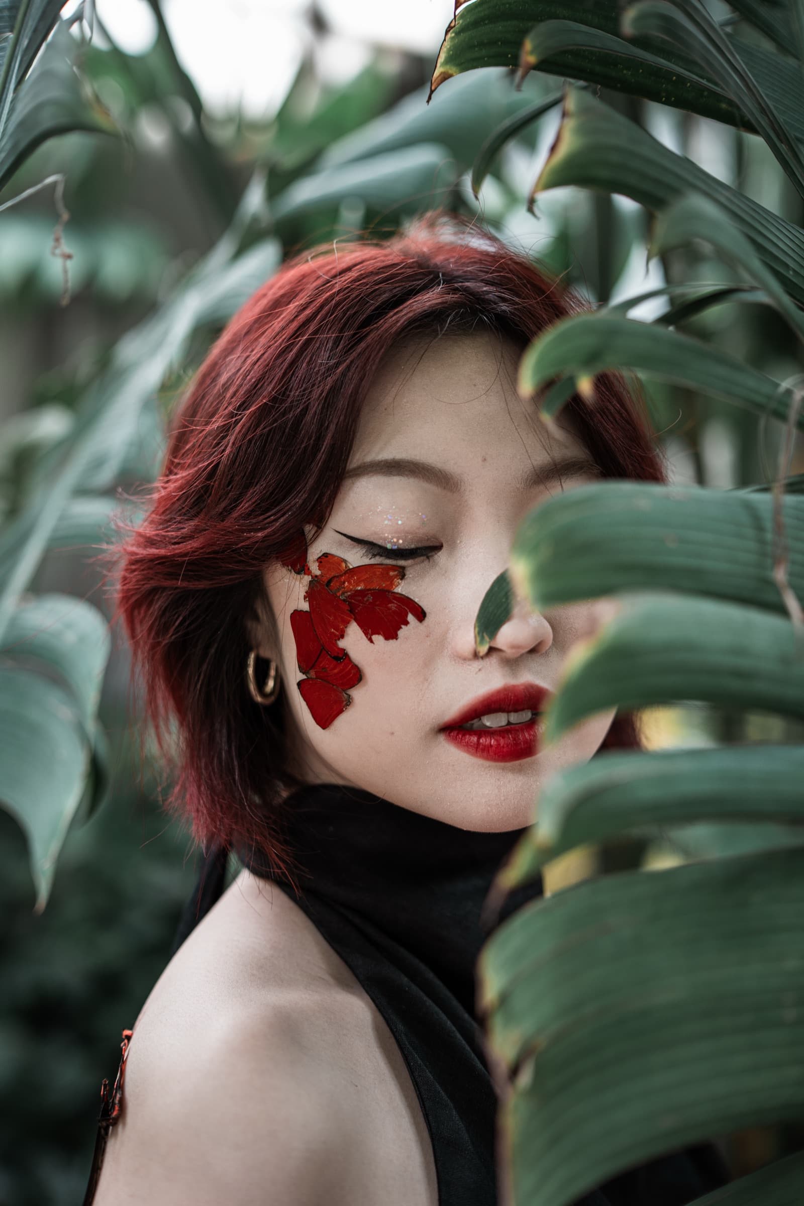 Eyes closed, wings layered across the cheekbone, red hair against palm fronds