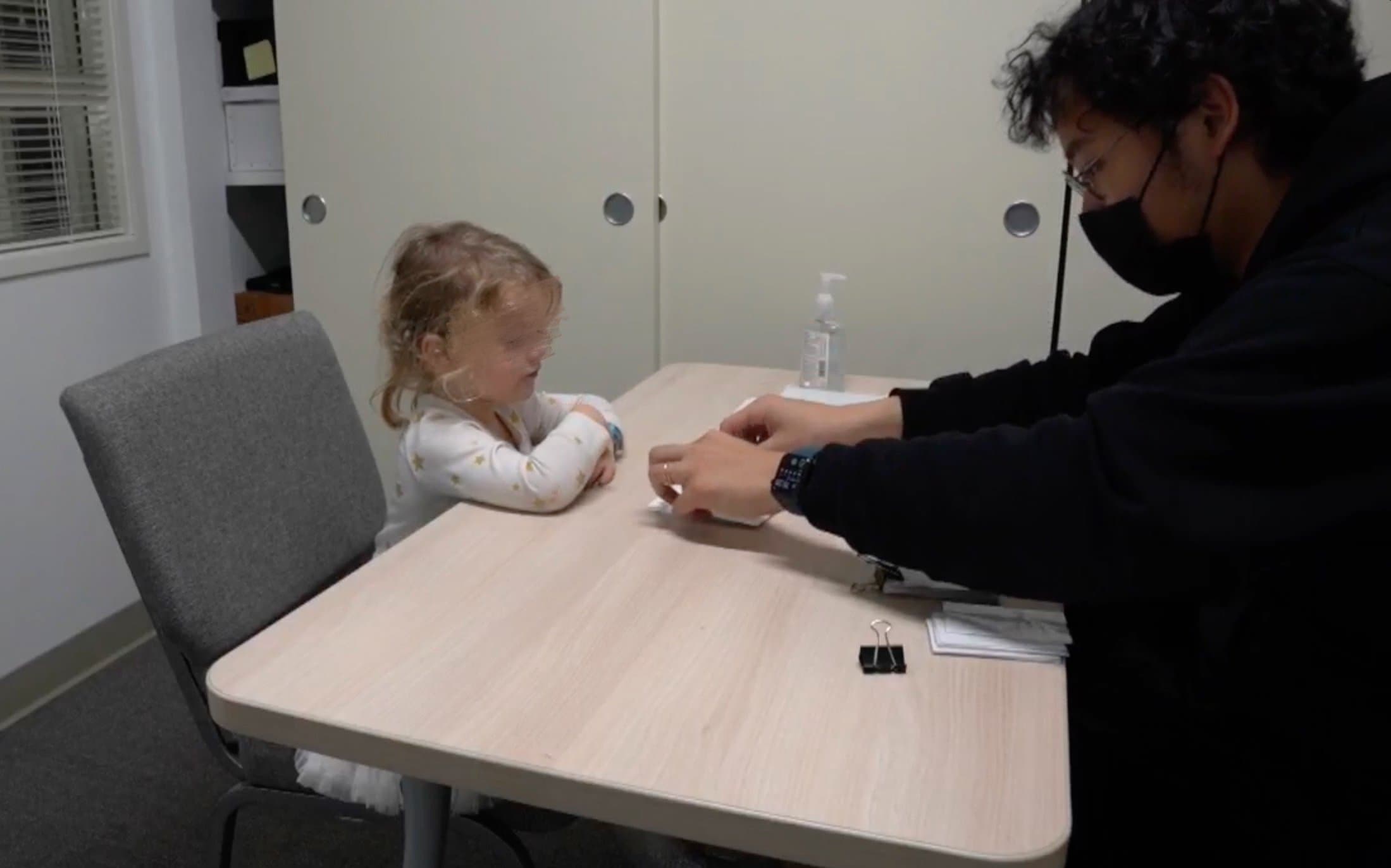An in-lab session: a masked researcher runs the card task across a small table from a young child